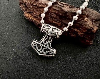 May include: A silver chain necklace with a silver Mjolnir pendant. The pendant has an intricate design with a detailed pattern.