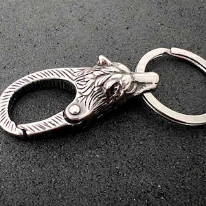 May include: A silver-colored keychain featuring a detailed wolf head design. The keychain includes a spring-loaded clasp and a split ring for attaching keys. The wolf head is intricately carved, showcasing fur and facial features.