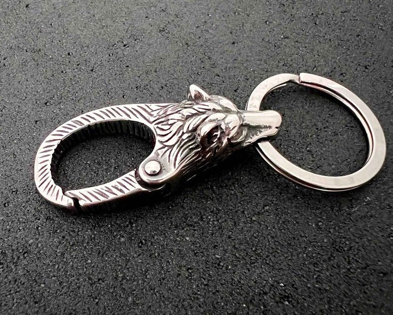 May include: A silver-colored keychain featuring a detailed wolf head design. The keychain includes a spring-loaded clasp and a split ring for attaching keys. The wolf head is intricately carved, showcasing fur and facial features.