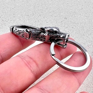 May include: A silver-colored keychain featuring a wolf head design. The wolf's head is detailed with sharp teeth and a fierce expression. The keychain includes a metal clasp and a circular key ring.