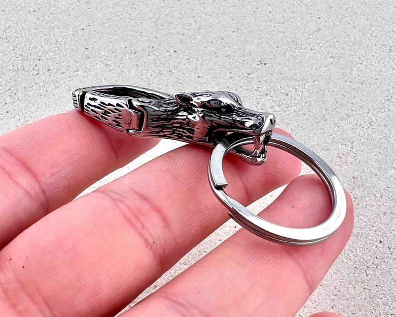 May include: A silver-colored keychain featuring a wolf head design. The wolf's head is detailed with sharp teeth and a fierce expression. The keychain includes a metal clasp and a circular key ring.