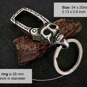 Puede incluir: Un llavero plateado con dise&ntilde;o de calavera y un anillo grande. El llavero mide 54 x 20 mm, y el anillo tiene un di&aacute;metro de 28 mm. El texto de la imagen dice "Size: 54 x 20mm 2.13 x 0.8 inch" y "The ring is 28 mm 1.1 inch in diameter."