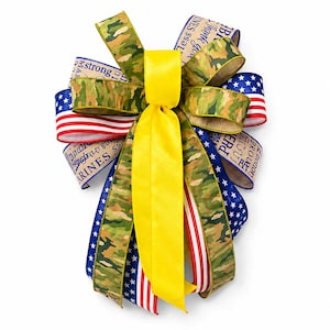 May include: A decorative bow featuring a combination of camouflage, American flag, and blue star-patterned ribbons. A bright yellow ribbon is centered, and ribbons with text that reads "Thank you" and "Marines" are also included.
