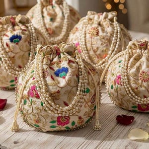 以下が含まれることがあります： ピンク、ブルー、グリーンの糸で施された複雑な花柄の刺繍とゴールドのアクセントが特徴の、オフホワイトの巾着袋のコレクション。各ポーチには、白いフェイクパールの多連ストランドで作られた装飾的なハンドルが付いています。