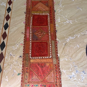 May include: A rectangular fabric wall hanging or table runner featuring a patchwork of red, orange, and gold embroidered panels. The piece is decorated with small white cowrie shells arranged in a border and along the edges, with small colorful tassels attached to the sides.