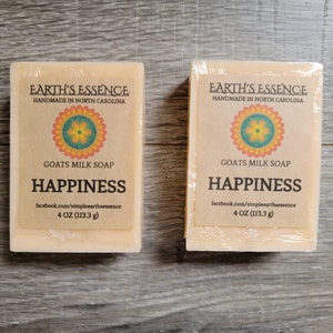 May include: Two bars of goat's milk soap, each wrapped in clear plastic. The labels read "Earth's Essence, Handmade in North Carolina," with a floral design and the word "HAPPINESS." Each bar weighs 4 oz (113.3 g).