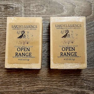 May include: Two bars of goat's milk soap, each wrapped in clear plastic. The labels read "Earth's Essence, Handmade in North Carolina" with a drawing and the words "Goats Milk Soap Open Range, 4 oz (113.3 g)."