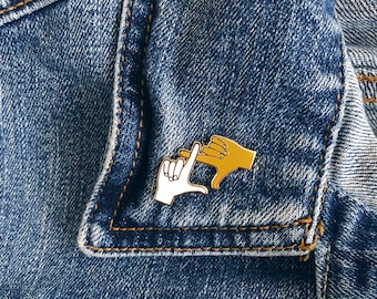 May include: A gold-colored enamel pin featuring two hands making a frame with their fingers. The hands are outlined in black and filled in with white and yellow enamel.