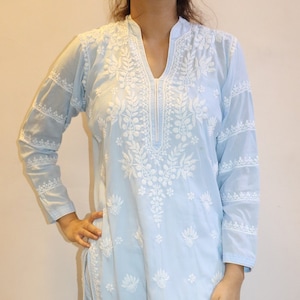 May include: A light blue embroidered kurta and white pants. The kurta features intricate white floral embroidery along the neckline, sleeves, and body. The pants are white and have a textured appearance. The outfit is worn with white sandals.
