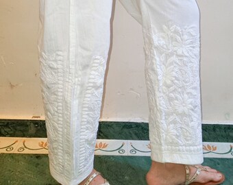 Può includere: Pantaloni bianchi in cotone con ricami bianchi intricati sulle gambe. I pantaloni sono indossati con sandali dorati.