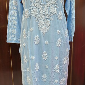 May include: A light blue tunic dress with intricate white floral embroidery. The long-sleeved dress features a mandarin collar and detailed patterns on the sleeves and bodice. The dress is displayed on a mannequin.