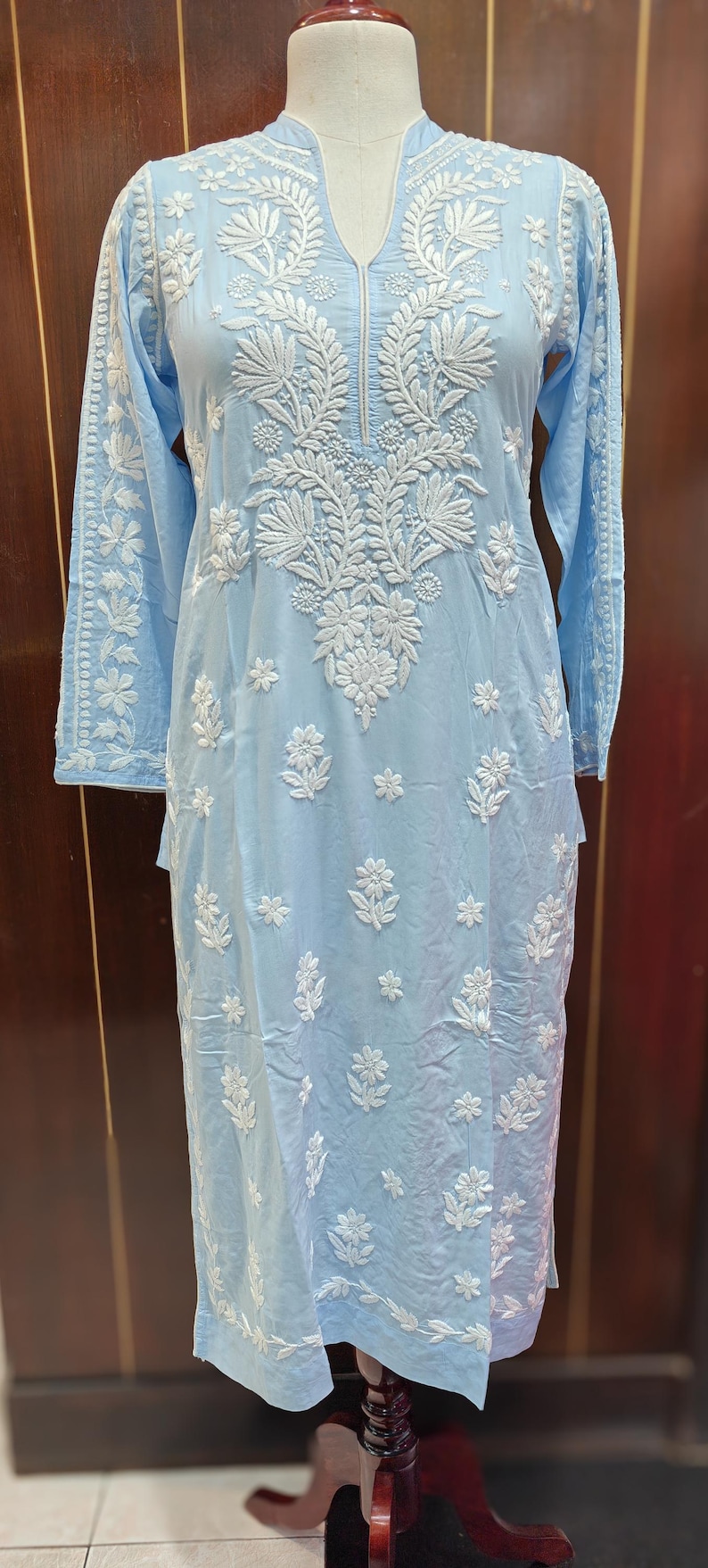 May include: A light blue tunic dress with intricate white floral embroidery. The long-sleeved dress features a mandarin collar and detailed patterns on the sleeves and bodice. The dress is displayed on a mannequin.