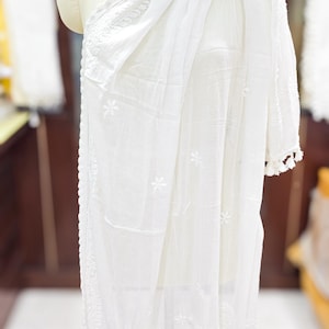 May include: A white, embroidered scarf and shawl draped over a mannequin. The scarf has a delicate floral pattern and tassel trim. The shawl is made of a lightweight fabric. The mannequin is on a dark wooden stand.