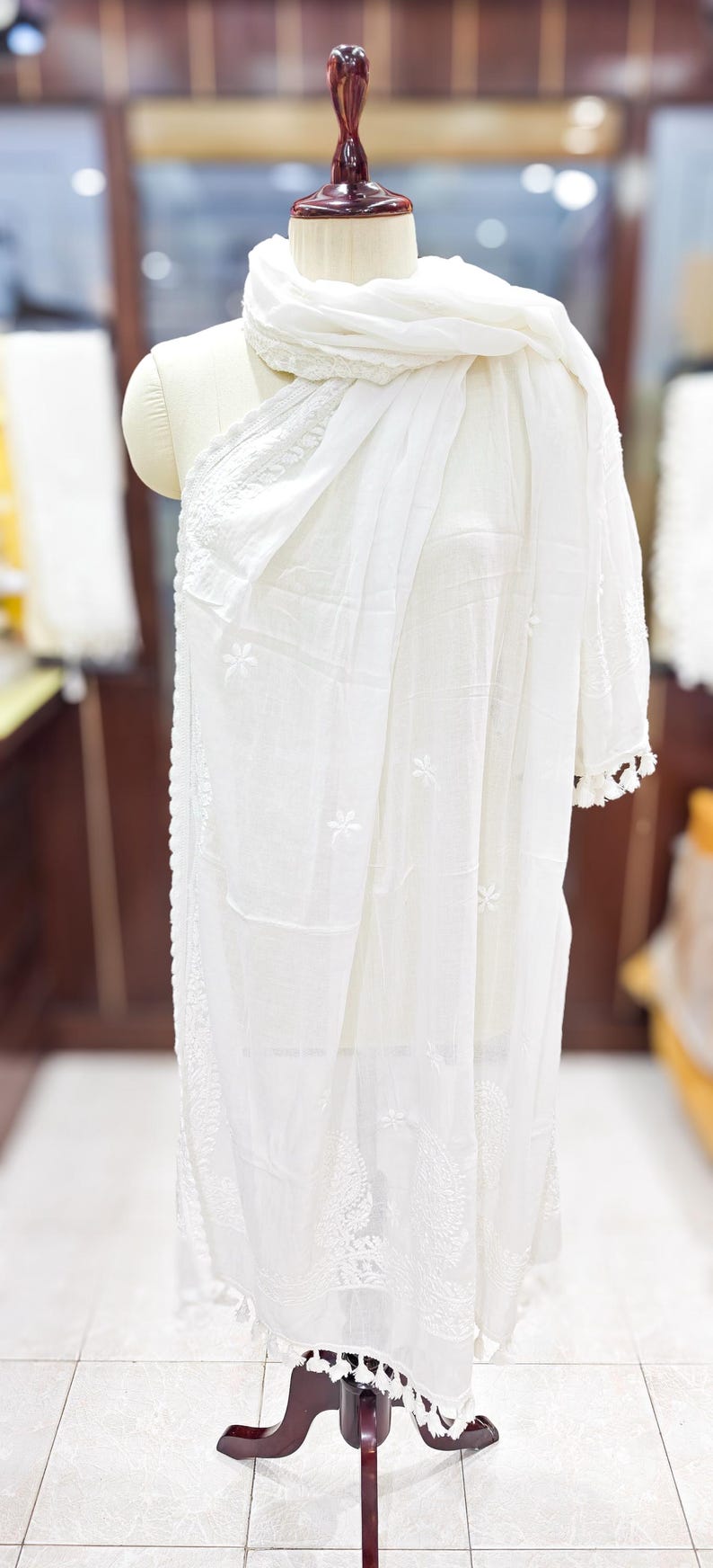 May include: A white, embroidered scarf and shawl draped over a mannequin. The scarf has a delicate floral pattern and tassel trim. The shawl is made of a lightweight fabric. The mannequin is on a dark wooden stand.