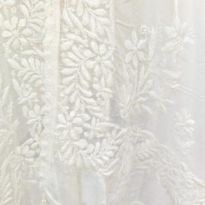 May include: Close-up of a white fabric with intricate floral and leaf embroidery. The design features detailed patterns of flowers and foliage, with a delicate fringe of tassels along the edge. The fabric appears to be lightweight and airy.