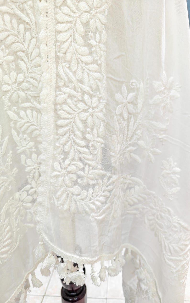 May include: Close-up of a white fabric with intricate floral and leaf embroidery. The design features detailed patterns of flowers and foliage, with a delicate fringe of tassels along the edge. The fabric appears to be lightweight and airy.