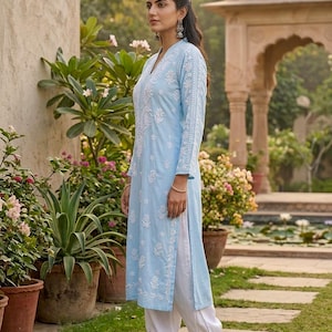 May include: A light blue tunic and white pants ensemble with intricate white embroidery. The long-sleeved tunic features a V-neck and side slits, paired with loose-fitting pants. The outfit is completed with gold sandals, creating a relaxed, elegant look.
