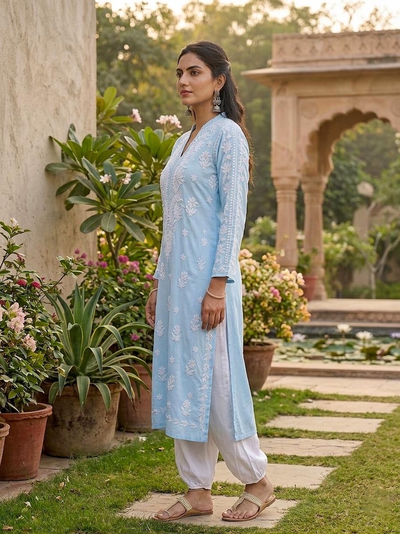 May include: A light blue tunic and white pants ensemble with intricate white embroidery. The long-sleeved tunic features a V-neck and side slits, paired with loose-fitting pants. The outfit is completed with gold sandals, creating a relaxed, elegant look.