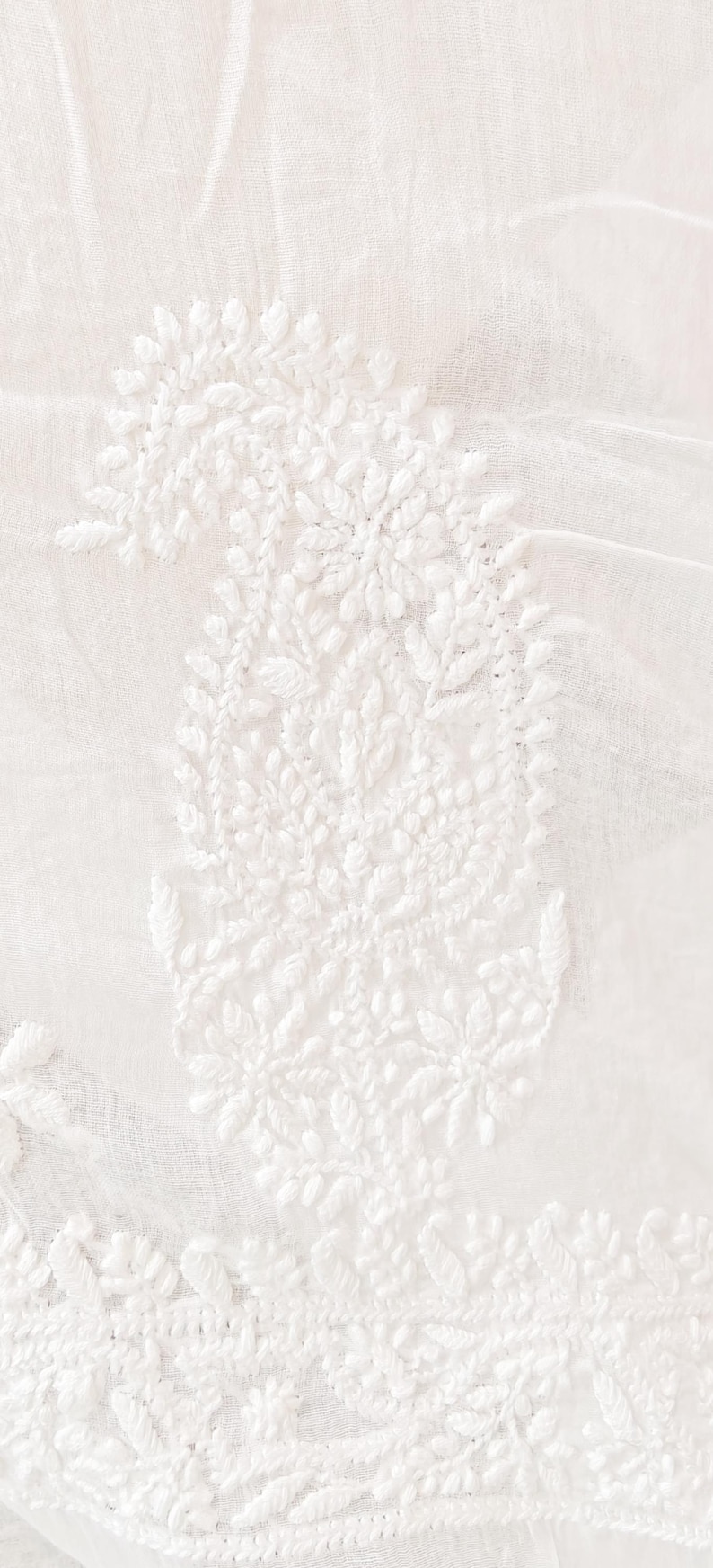 May include: Close-up of white fabric with intricate, raised embroidery. The design features a large paisley shape with floral and leaf details. The fabric has a slightly sheer quality, and the embroidery is densely packed.