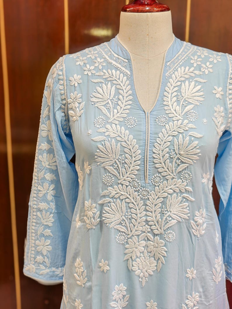 May include: A light blue tunic with intricate white floral embroidery. The long-sleeved top features a mandarin collar and a V-neckline. The white embroidery is detailed with floral and leaf patterns, creating a delicate and elegant design.