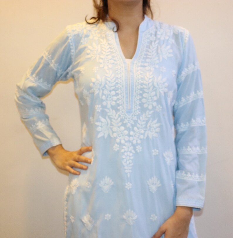 May include: A light blue tunic with long sleeves and intricate white floral embroidery. The tunic features a mandarin collar and a V-neckline. The embroidery is concentrated around the neckline and extends down the front, with additional detailing on the sleeves.