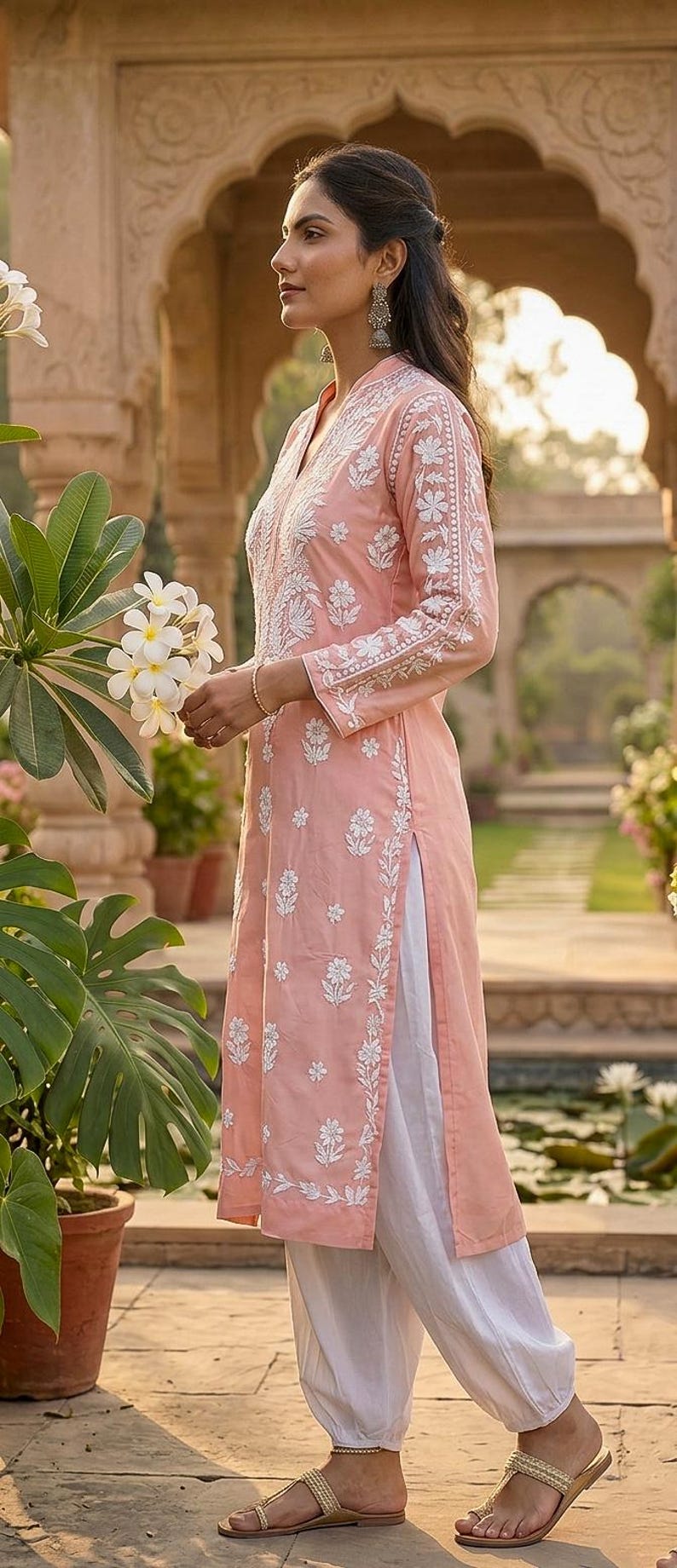 Puede incluir: Conjunto de t&uacute;nica melocot&oacute;n claro y pantal&oacute;n blanco con intrincado bordado floral blanco. La t&uacute;nica de manga larga presenta un cuello en V y aberturas laterales. El atuendo se combina con sandalias doradas.