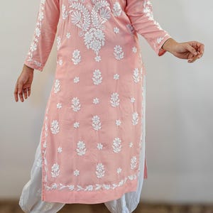 Puede incluir: Una t&uacute;nica rosa claro con bordado floral blanco. La t&uacute;nica de manga larga presenta un cuello mandar&iacute;n y una abertura en el dobladillo. La t&uacute;nica se combina con pantalones blancos y sandalias.