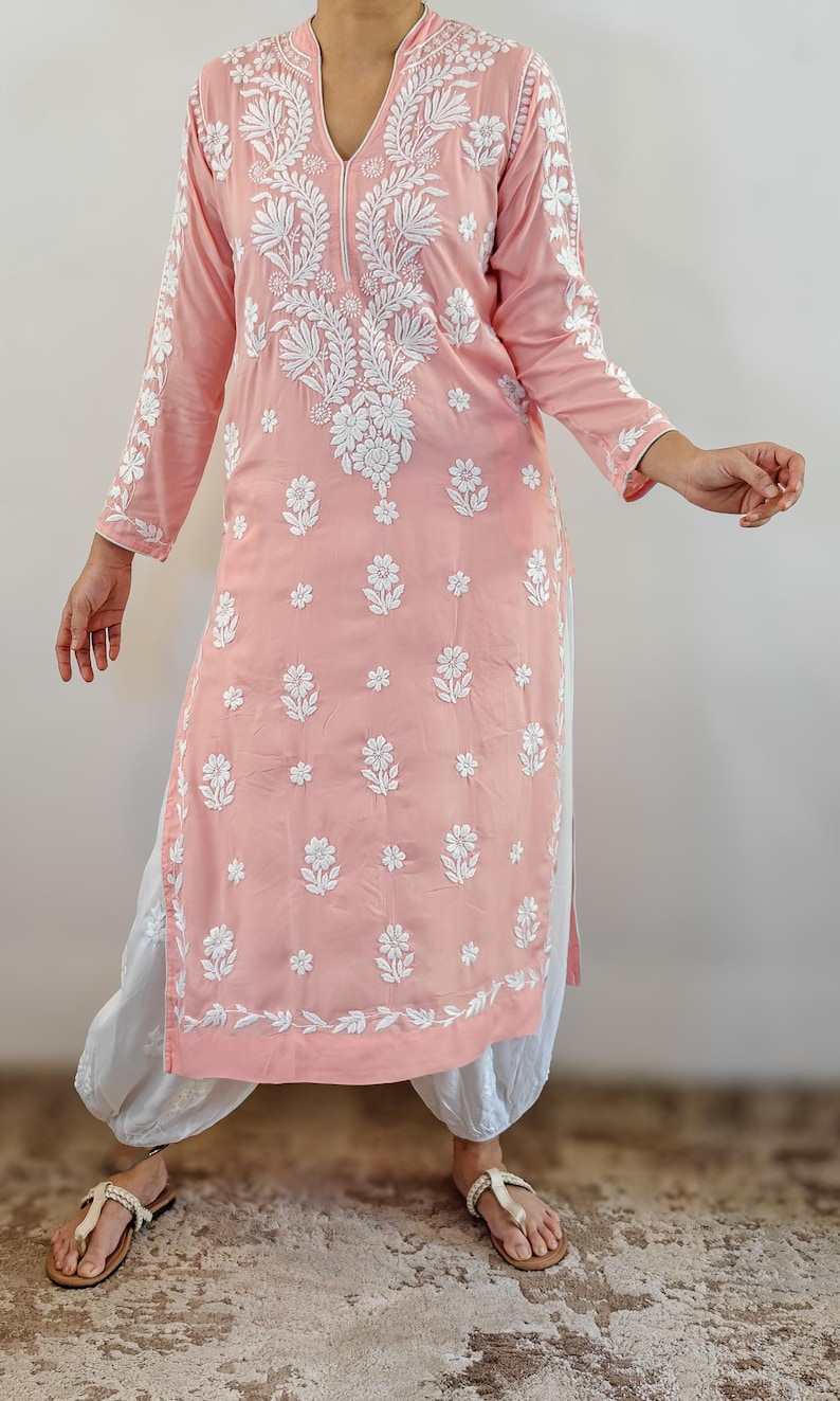 Puede incluir: Una t&uacute;nica rosa claro con bordado floral blanco. La t&uacute;nica de manga larga presenta un cuello mandar&iacute;n y una abertura en el dobladillo. La t&uacute;nica se combina con pantalones blancos y sandalias.