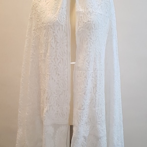 May include: White embroidered shawl with a delicate floral pattern. The shawl is made of a sheer fabric and has a flowing drape.