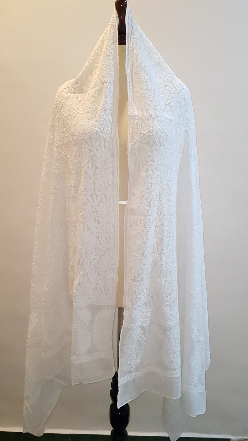May include: White embroidered shawl with a delicate floral pattern. The shawl is made of a sheer fabric and has a flowing drape.