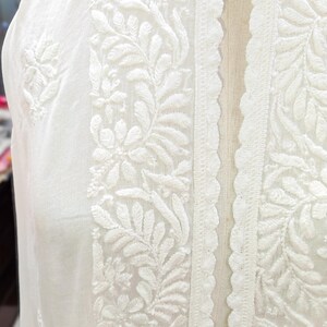 May include: Close-up of a white garment with intricate floral embroidery. The design features detailed leaf and flower patterns along the edges and center, with a scalloped lace trim. The fabric appears to be lightweight.