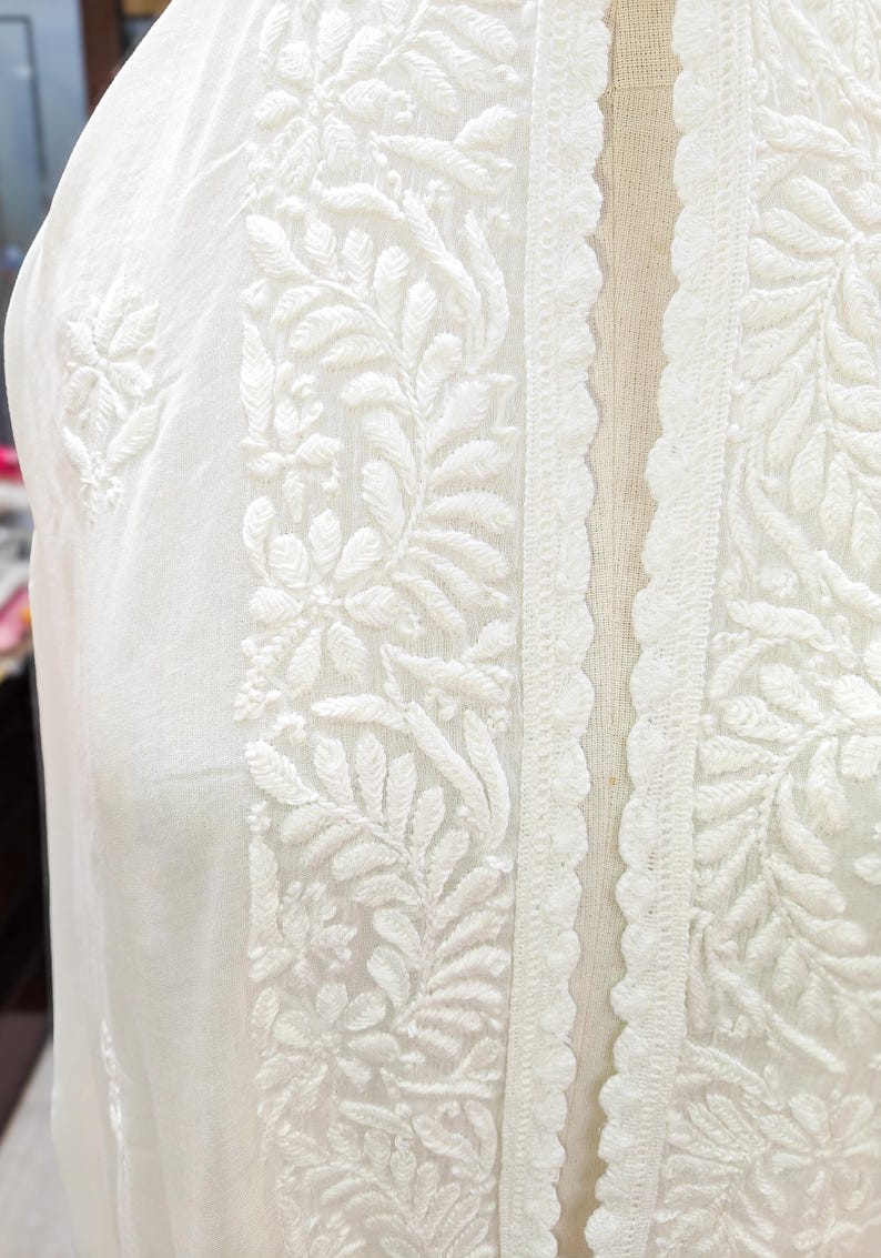 May include: Close-up of a white garment with intricate floral embroidery. The design features detailed leaf and flower patterns along the edges and center, with a scalloped lace trim. The fabric appears to be lightweight.