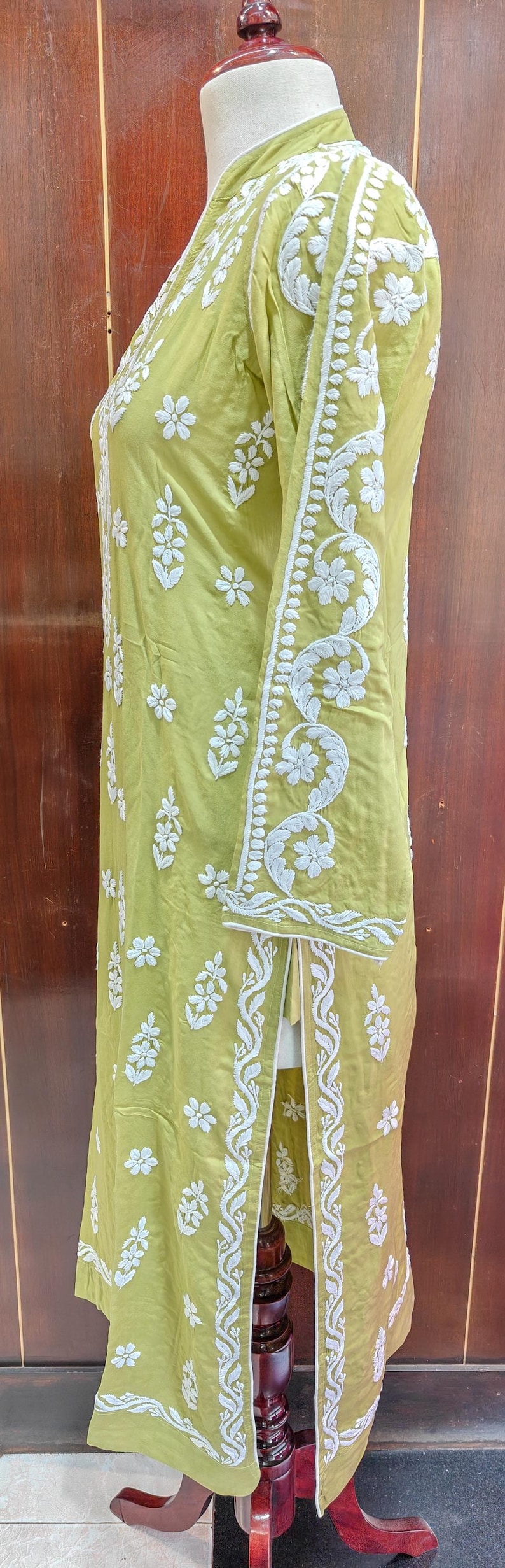 Puede incluir: Una t&uacute;nica verde claro con intrincados bordados florales blancos. La prenda de manga larga presenta patrones detallados a lo largo de las mangas y los bordes, con una abertura en el dobladillo. La t&uacute;nica se exhibe en un maniqu&iacute;.