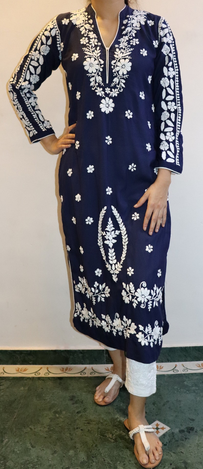 May include: Navy blue tunic dress with white floral embroidery. The long-sleeved dress features a mandarin collar and a split neckline. White floral embroidery adorns the sleeves, bodice, and hem. The dress is paired with white pants.