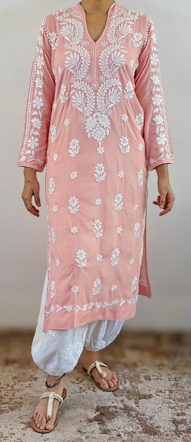 Puede incluir: Una t&uacute;nica rosa claro con intrincados bordados florales blancos. La t&uacute;nica tiene cuello en V y mangas tres cuartos. El modelo tambi&eacute;n lleva pantalones blancos y sandalias.