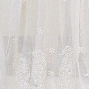May include: White embroidered fabric with floral and paisley patterns. The fabric has a sheer quality, with detailed stitching and a fringe of tassels along the bottom edge. The design is delicate and intricate.