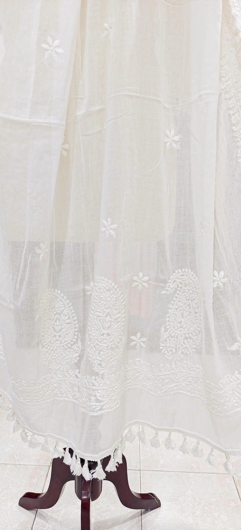 May include: White embroidered fabric with floral and paisley patterns. The fabric has a sheer quality, with detailed stitching and a fringe of tassels along the bottom edge. The design is delicate and intricate.