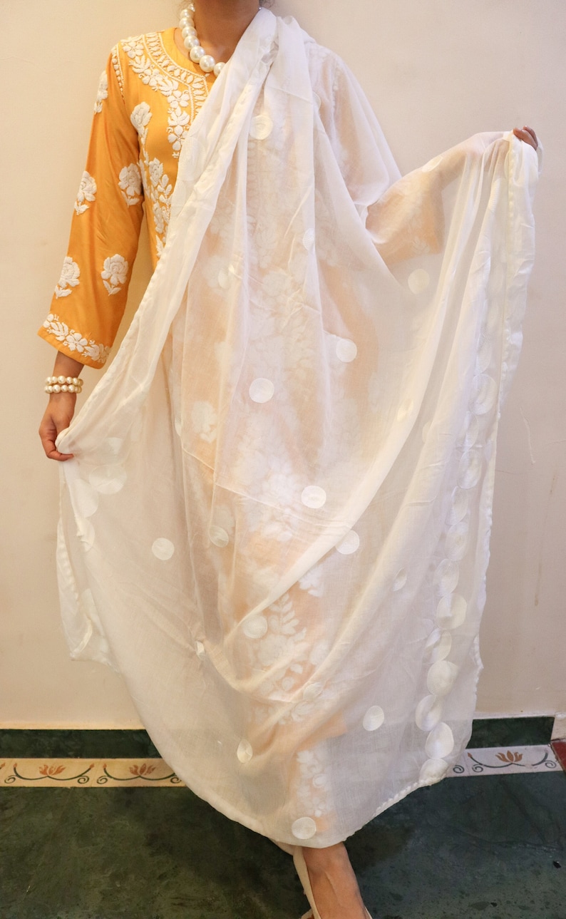 May include: A white sheer scarf with a polka dot pattern. The scarf is draped over a person wearing a yellow embroidered top.