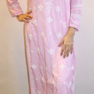 Puede incluir: Un vestido tipo t&uacute;nica rosa claro con mangas largas y un intrincado bordado floral blanco. El vestido presenta un cuello en V y se combina con pantalones blancos. El atuendo es adecuado para ropa casual.