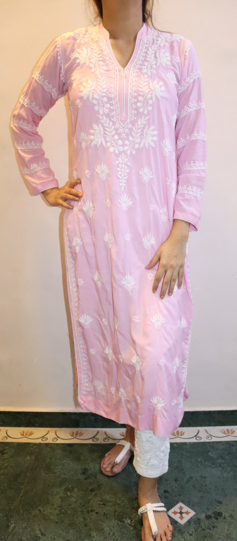 Puede incluir: Un vestido tipo t&uacute;nica rosa claro con mangas largas y un intrincado bordado floral blanco. El vestido presenta un cuello en V y se combina con pantalones blancos. El atuendo es adecuado para ropa casual.