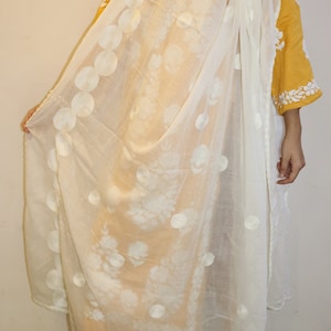 May include: A white sheer scarf with a floral pattern and white embroidered circles. The scarf is draped over a person wearing a yellow embroidered top.