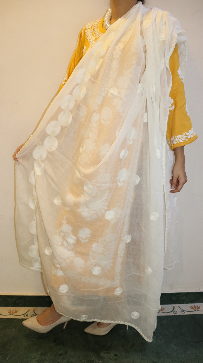 May include: A white sheer scarf with a floral pattern and white embroidered circles. The scarf is draped over a person wearing a yellow embroidered top.