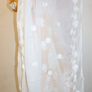 May include: White sheer scarf with embroidered white circles. The scarf is draped over a yellow and white outfit.