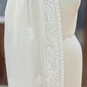 May include: A white, sheer scarf with intricate floral embroidery along the edges and scattered across the fabric. The scarf is draped over a mannequin, showcasing its delicate texture and design.