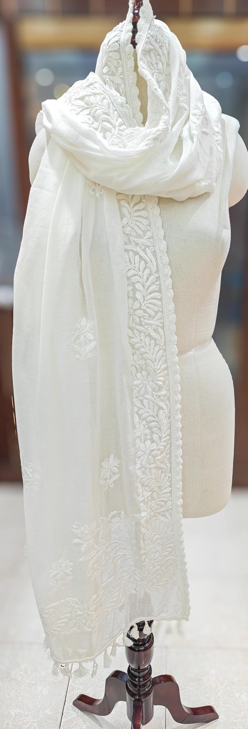May include: A white, sheer scarf with intricate floral embroidery along the edges and scattered across the fabric. The scarf is draped over a mannequin, showcasing its delicate texture and design.