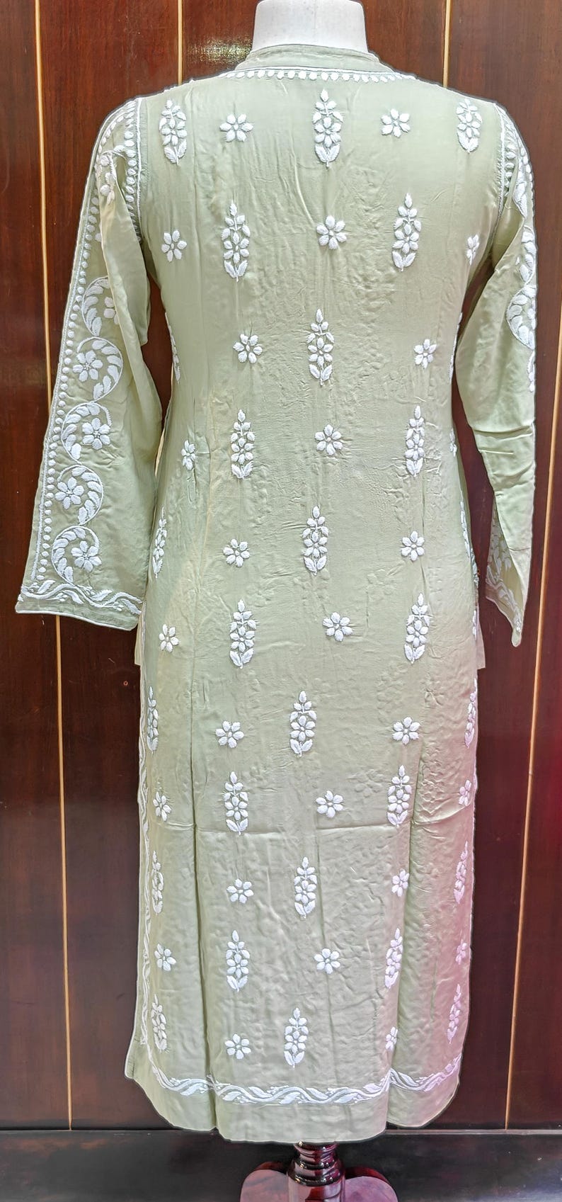 May include: A light green tunic dress with long sleeves and a mandarin collar. The dress is adorned with white floral embroidery. The embroidery is concentrated on the sleeves, front, and back of the dress. The dress is made of a lightweight fabric.