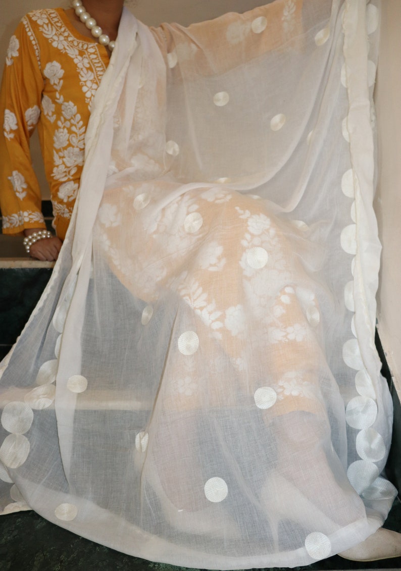 May include: White sheer scarf with embroidered white circles and floral designs. The scarf is draped over a yellow embroidered top.