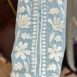 May include: Close-up of a light blue fabric with intricate white floral embroidery. The design features large flowers and delicate leaves, with a vertical line of small white stitches down the center. The fabric appears to be a lightweight material.