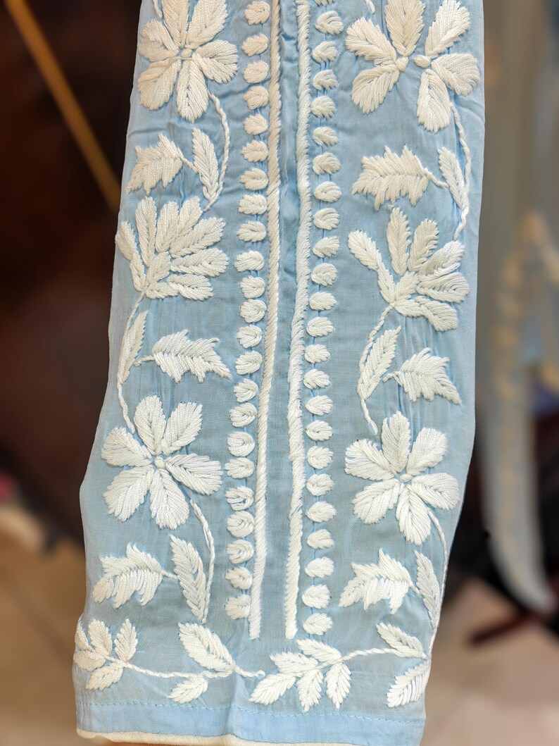 May include: Close-up of a light blue fabric with intricate white floral embroidery. The design features large flowers and delicate leaves, with a vertical line of small white stitches down the center. The fabric appears to be a lightweight material.