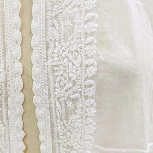 May include: Close-up of white fabric with intricate embroidery. The design features floral and leaf patterns along the edges and center. The fabric has a delicate, sheer appearance, suggesting a lightweight garment or textile.